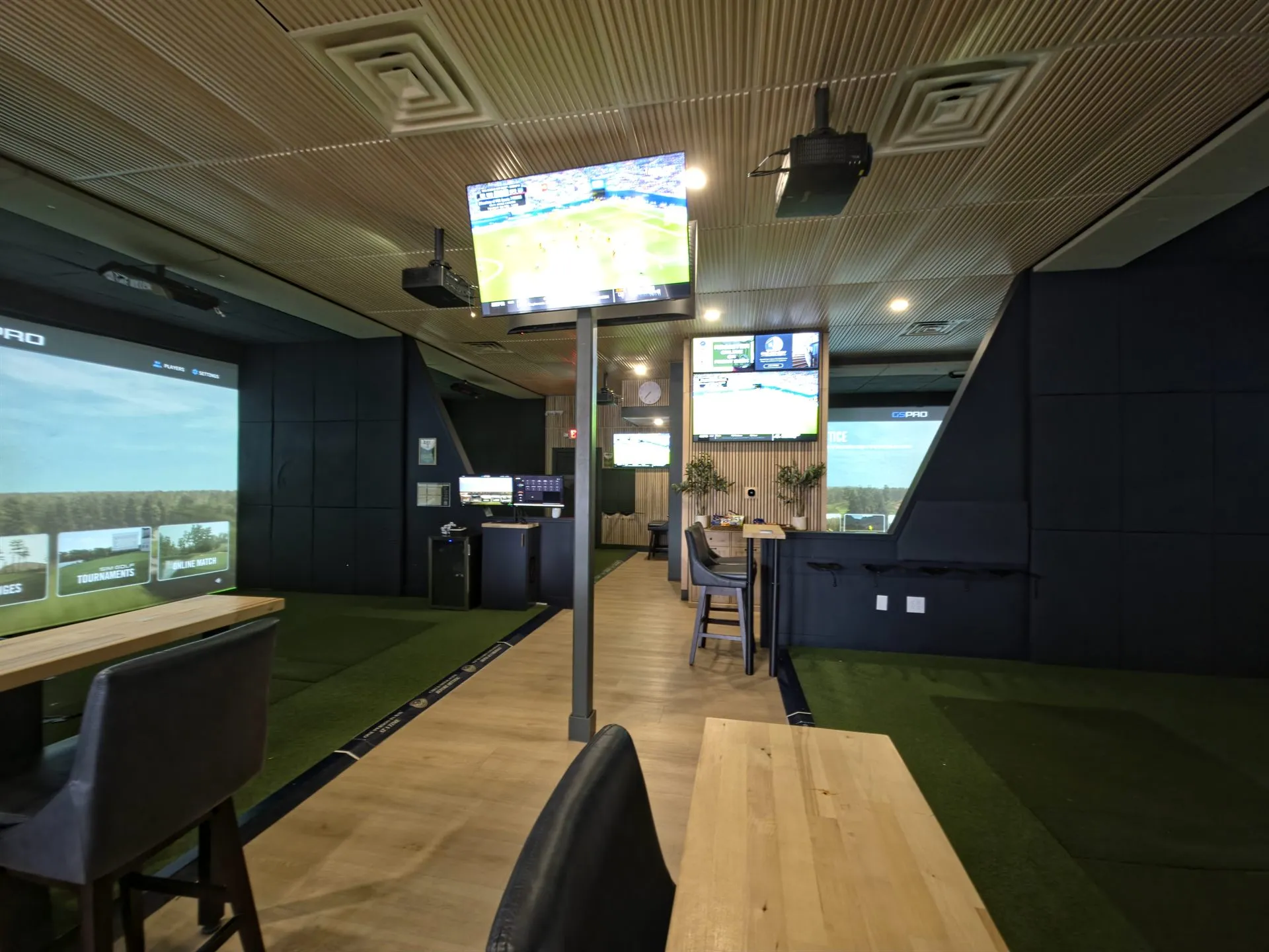 Row of golf simulator bays with green turf hitting lanes and overhead sports TVs at All Seasons, Kansas City