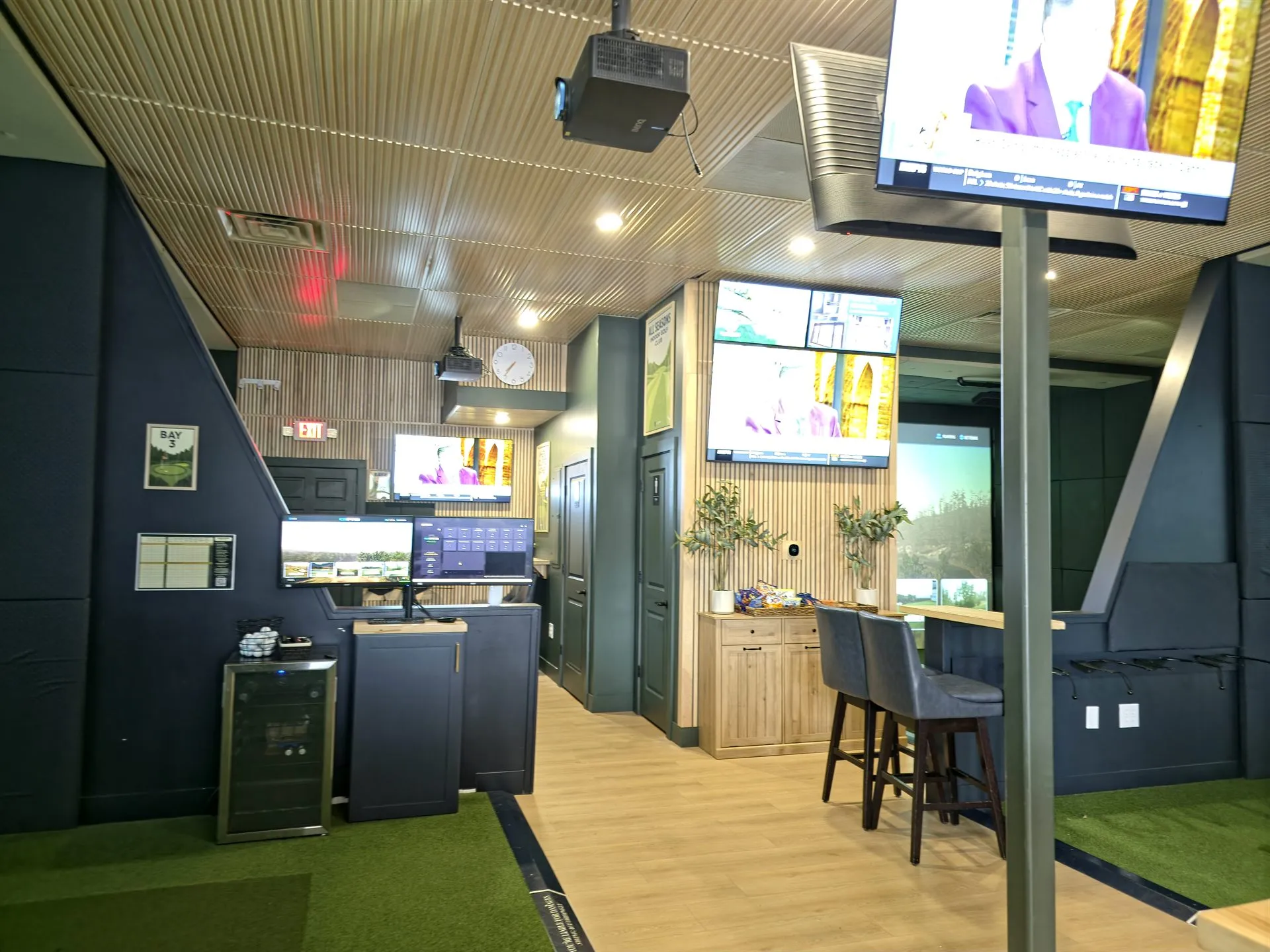 Indoor golf lounge with sports TVs, a beverage fridge, and bar seating at All Seasons, Kansas City